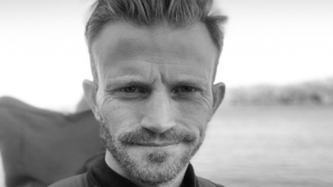 A close up Andy Caine face. He is wearing a rucksack training for his walk from John O’Groats to Land’s End. There are fields are trees blurred in the background.